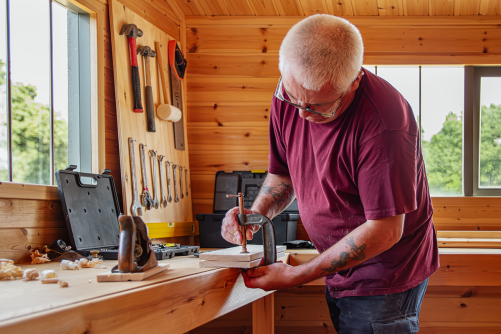 Superior Shed Workshop Man securing a piece of timber with a clamp on a workshop bench, surrounded by woodworking tools.